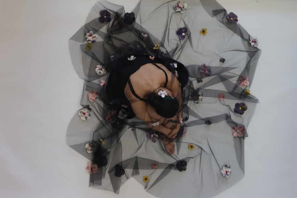 Person wearing a black dress stands behind and beneath a sheer black veil decorated with colorful fabric flowers, partially obscuring their face and body.