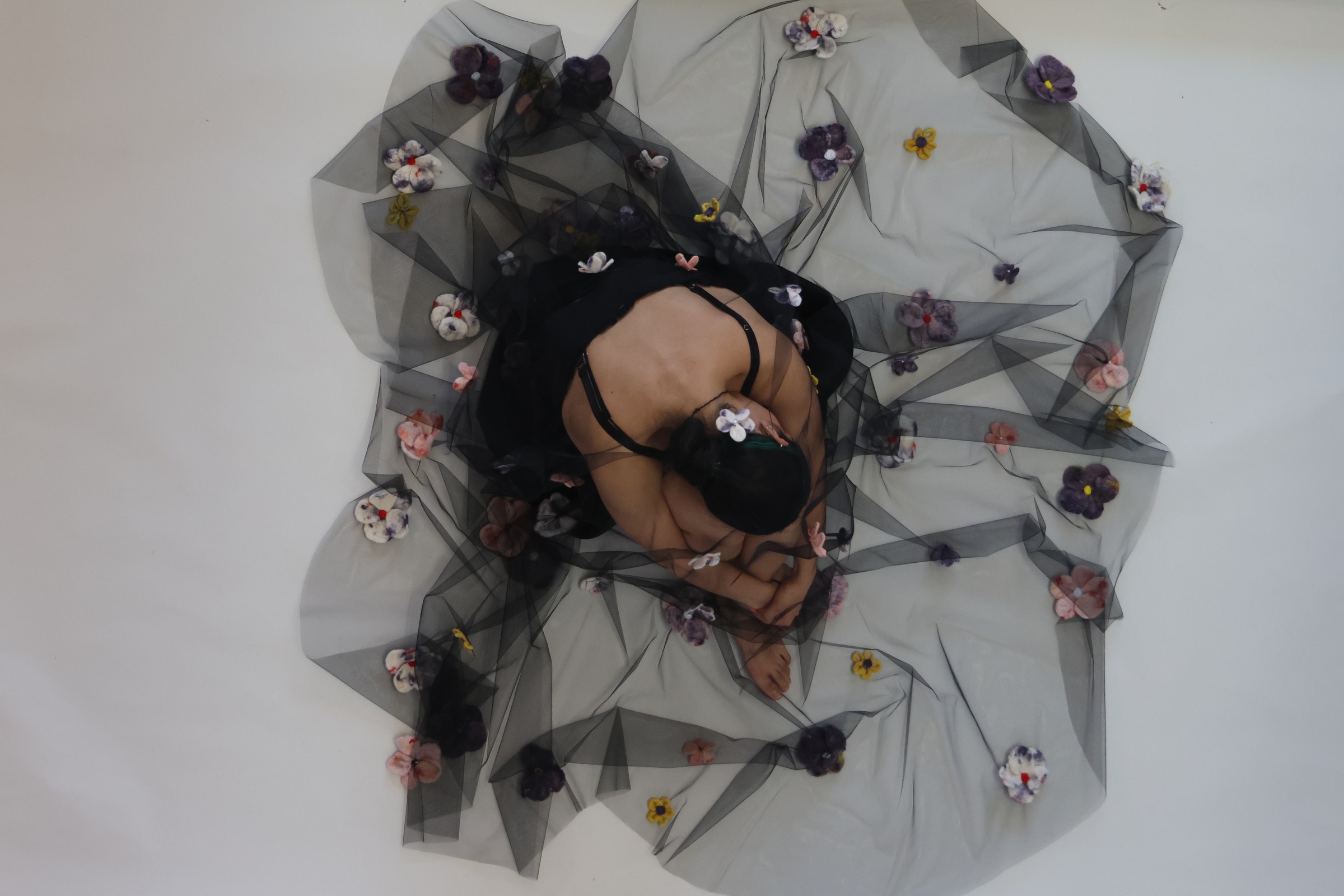 Person wearing a black dress stands behind and beneath a sheer black veil decorated with colorful fabric flowers, partially obscuring their face and body.