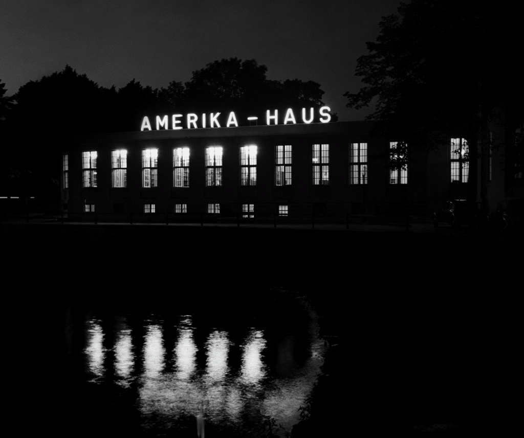 A nighttime photo of the Amerika-Haus building with illuminated windows and its name glowing on the rooftop, reflecting on the dark water below.