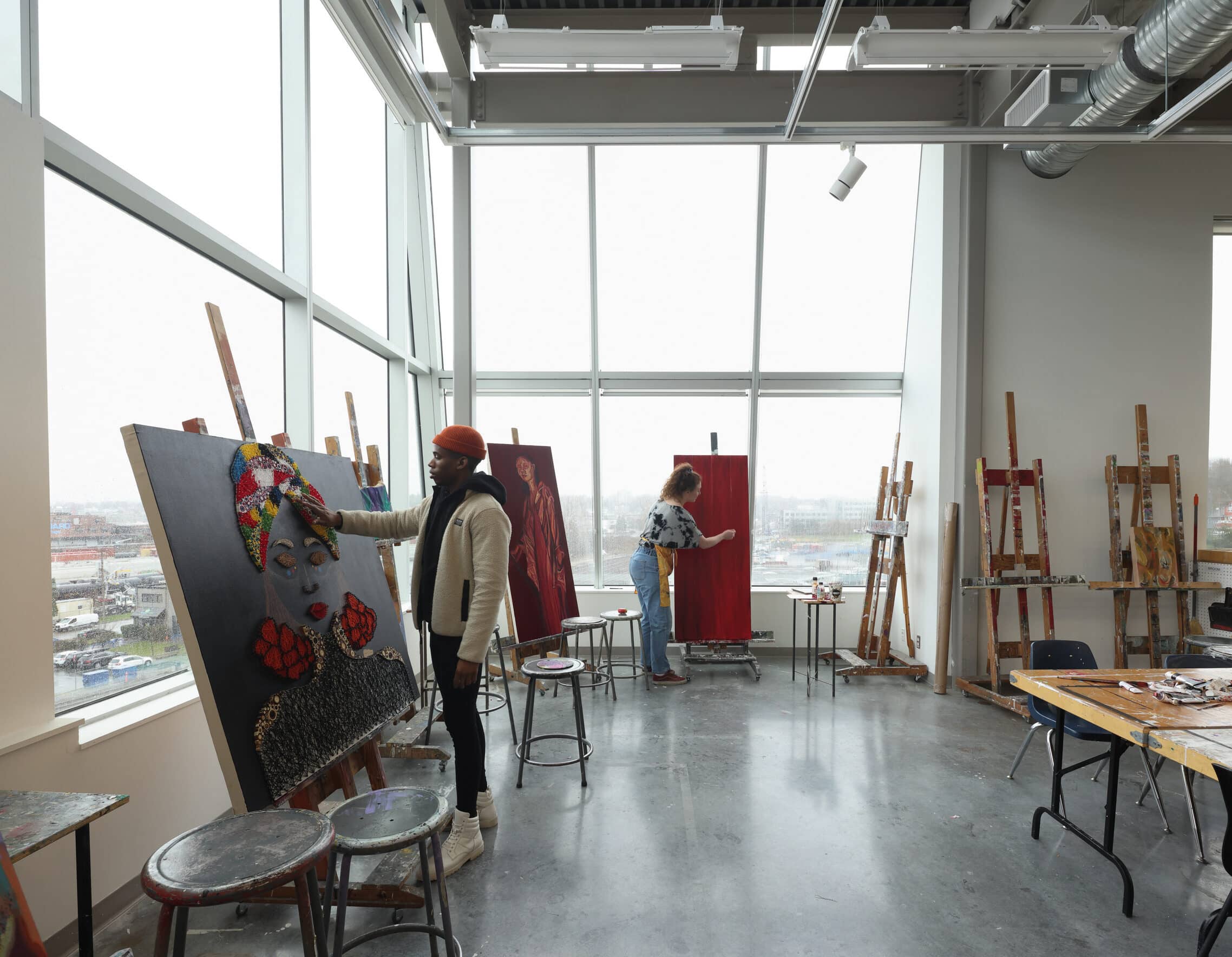 Students painting large canvases in a bright art studio with tall windows and easels.