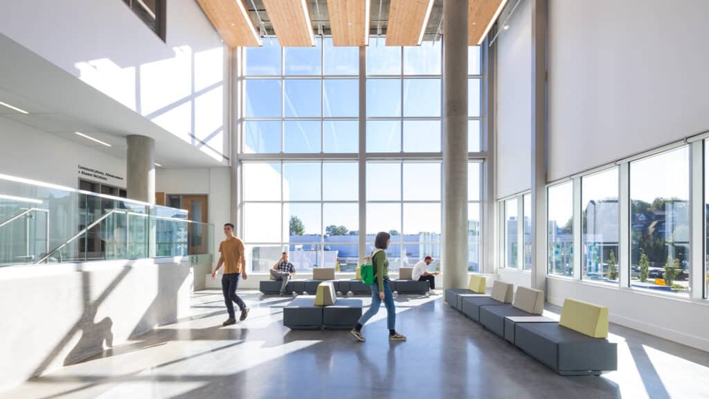 Spacious, sunlit lobby with high ceilings, large floor-to-ceiling windows, and wood-accented ceiling panels. People walk and sit on modern gray and green modular seating, with polished concrete floors and glass railings adding to the open, airy feel.