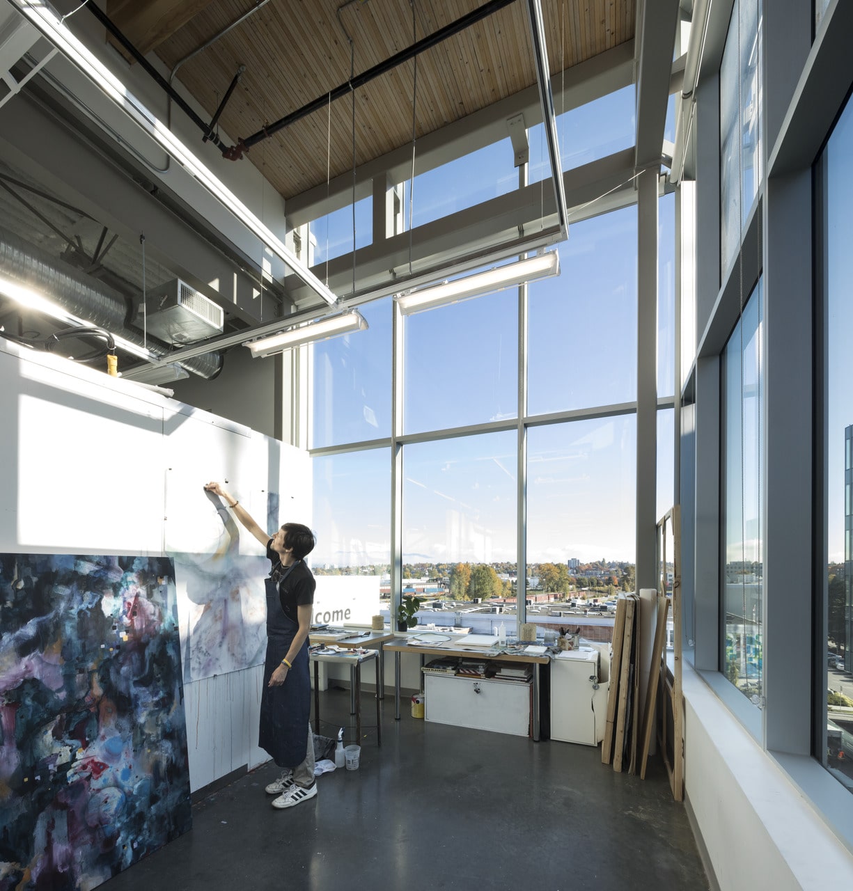 An artist works on a large abstract painting in a bright studio with tall windows overlooking the city.