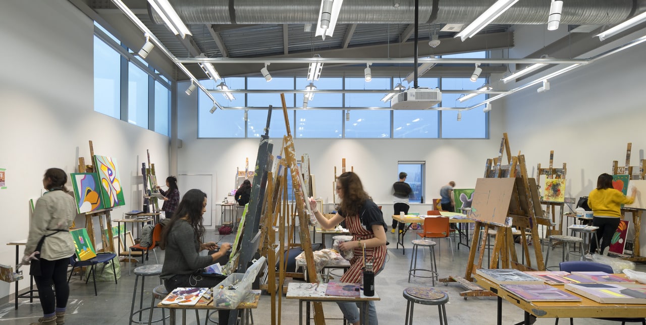 Art students working at easels in a bright studio classroom with large windows, painting and drawing colorful works on canvas.