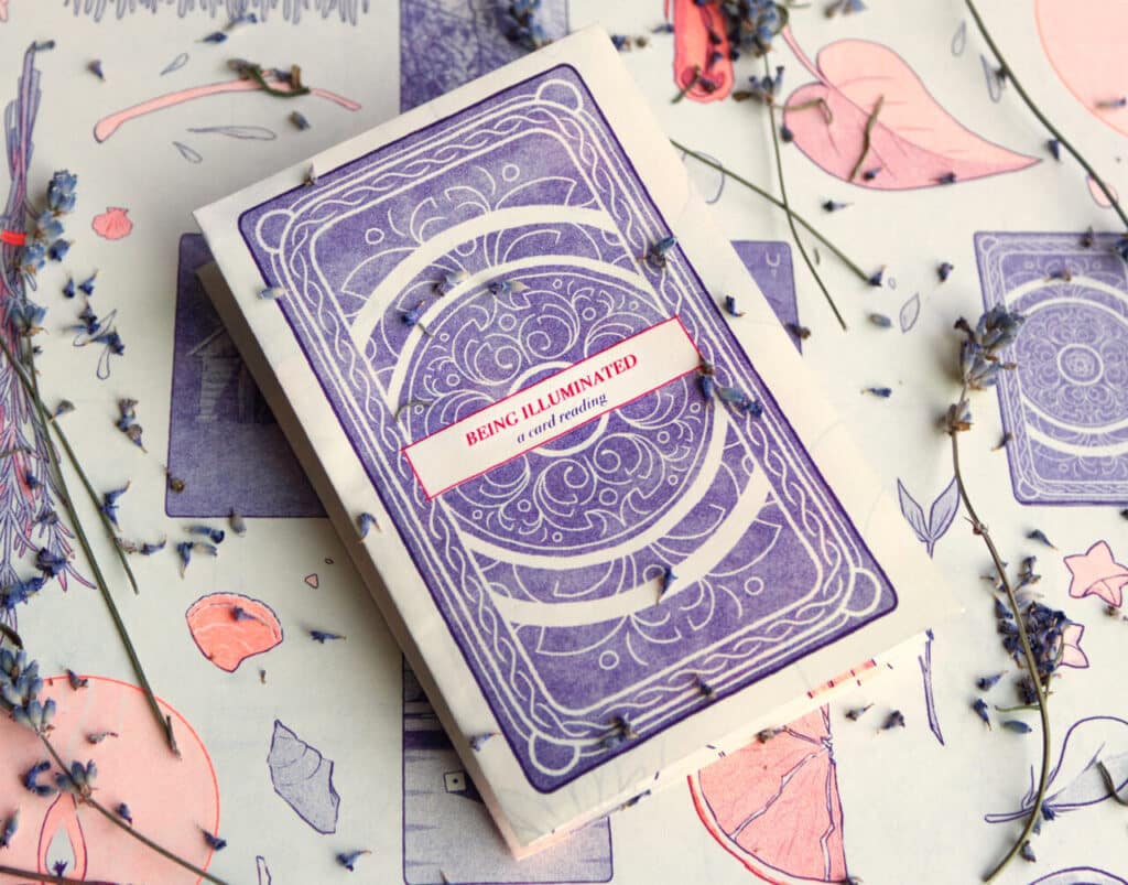 A handmade booklet with a purple illustrated cover titled Being Illuminated: a card reading, surrounded by dried flowers and illustrated paper.