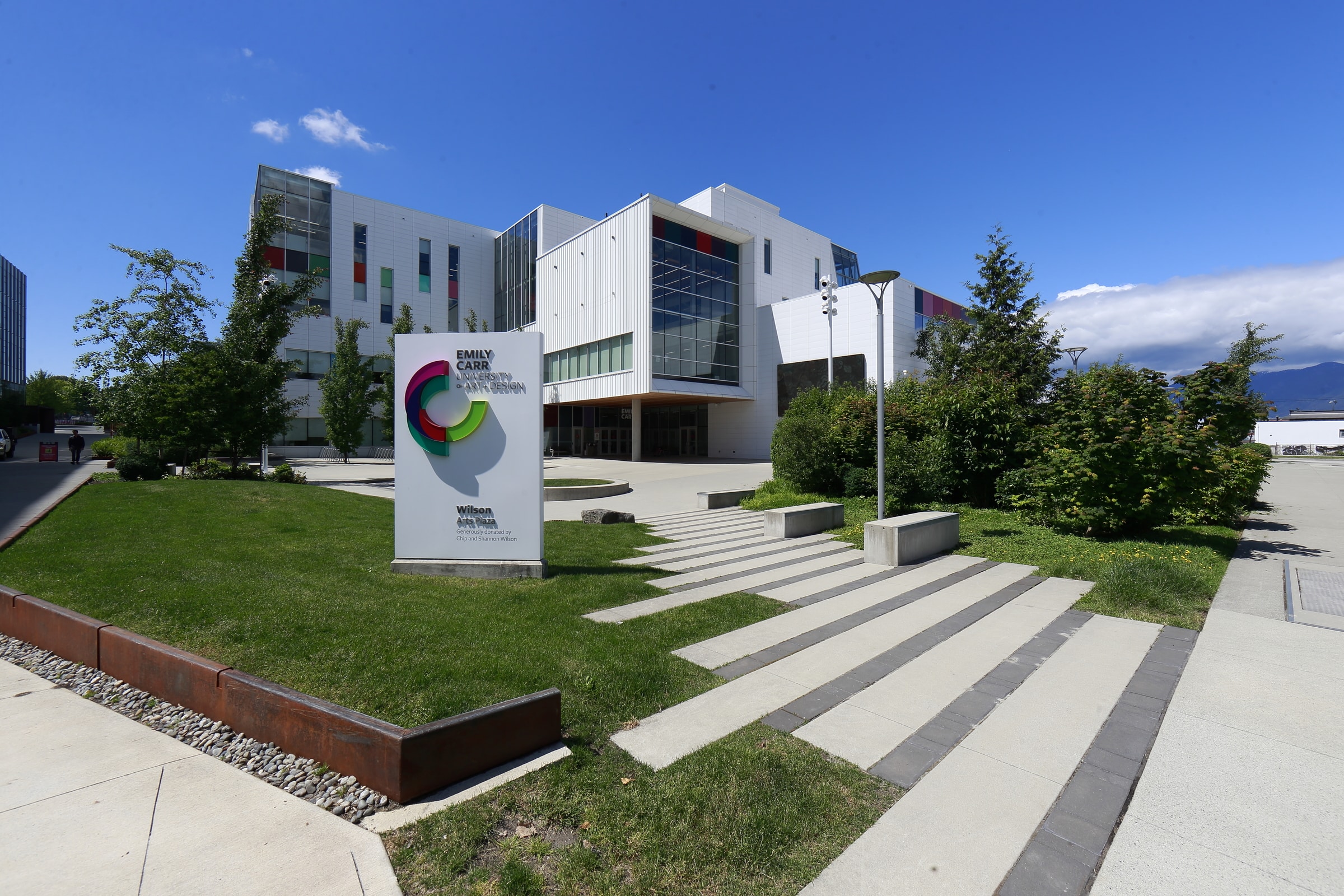 Exterior view of Emily Carr University of Art and Design on a sunny day, showing the modern white building with large windows and colorful accents, landscaped green lawn, and a prominent campus sign in the foreground.