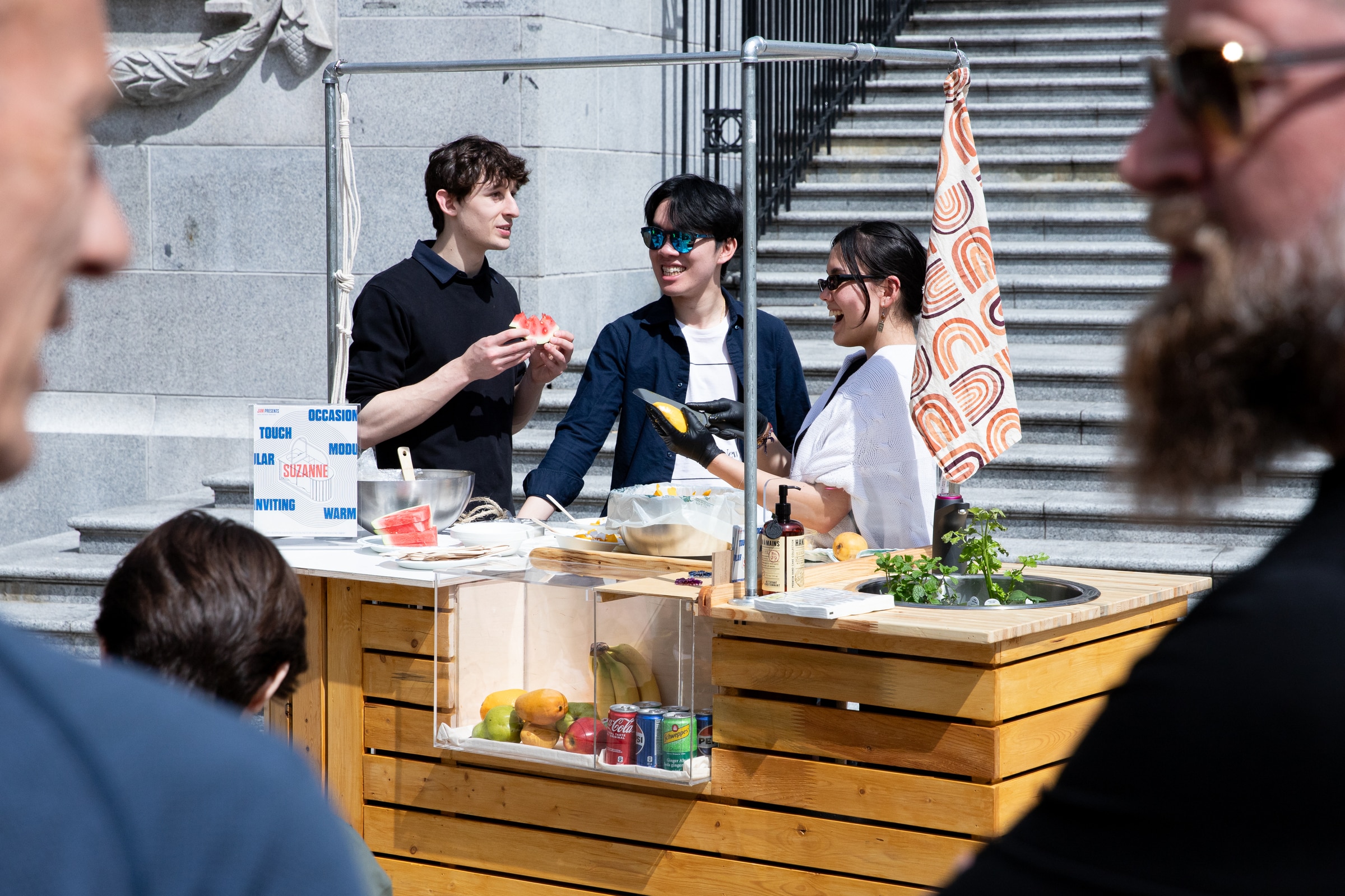 Three people smiling and chatting around a wooden food cart with fruit, drinks, and kitchen tools displayed, set outdoors in front of stone steps on a sunny day.