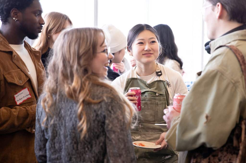 Students gather in conversation at a campus event, holding drinks and snacks while socializing in a bright indoor space.