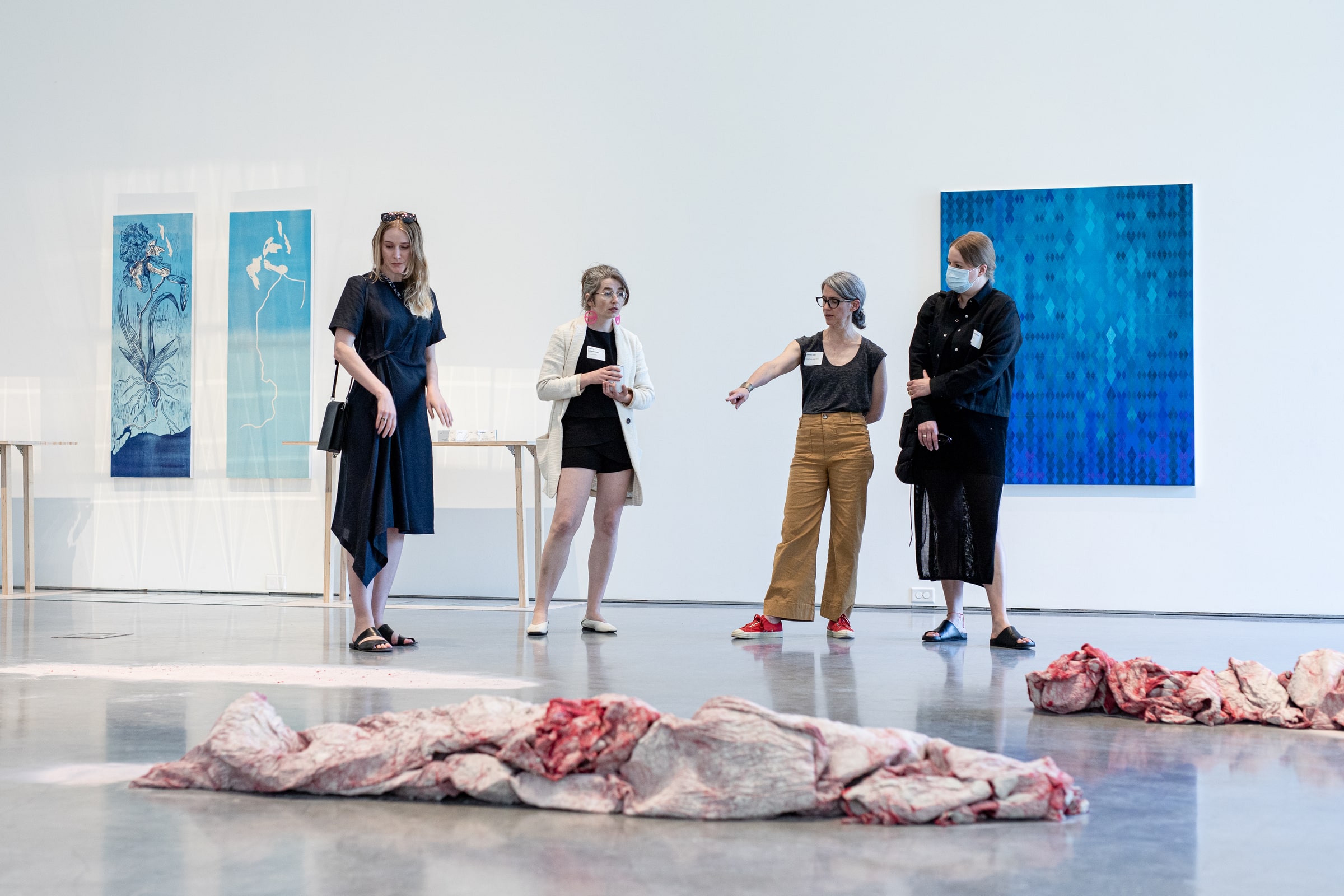 Four people stand in a gallery discussing an installation of fabric forms on the floor, with large blue-toned paintings and textile works displayed on the wall behind them.