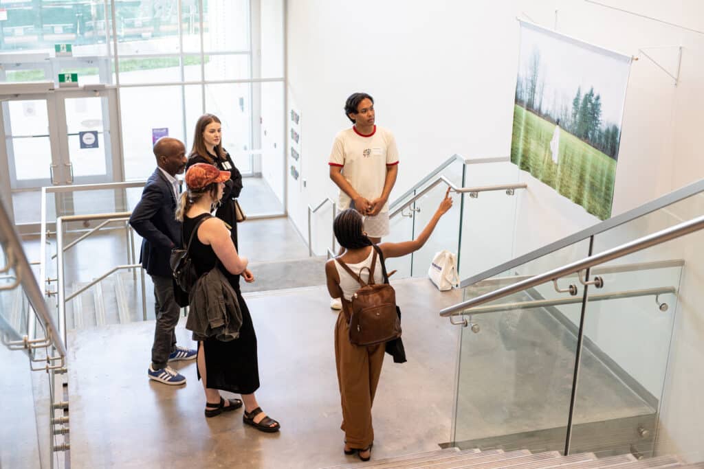 Five people view an artwork on a wall. The wall is located along a stairwell. The camera is at the top of the stairs; we are looking down at the people.