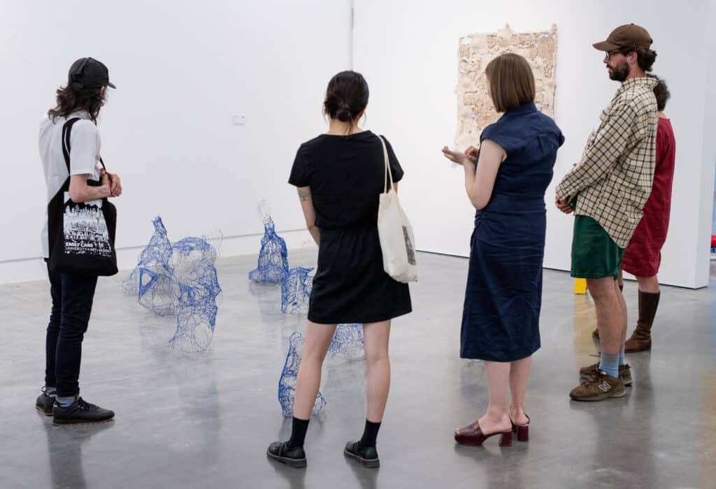 A group of five people stand in a gallery observing a floor installation of abstract blue wire sculptures. A textured rectangular artwork is mounted on the white wall behind them.