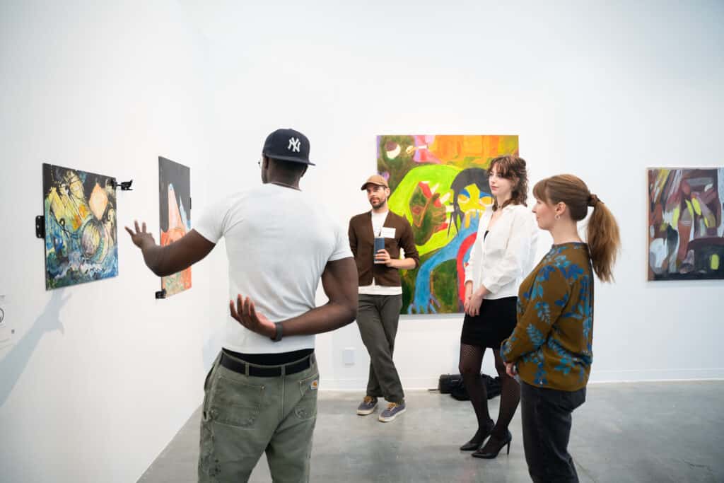 Group of students engaged in a critique session in front of colorful paintings, one student gesturing toward the work while others listen attentively.