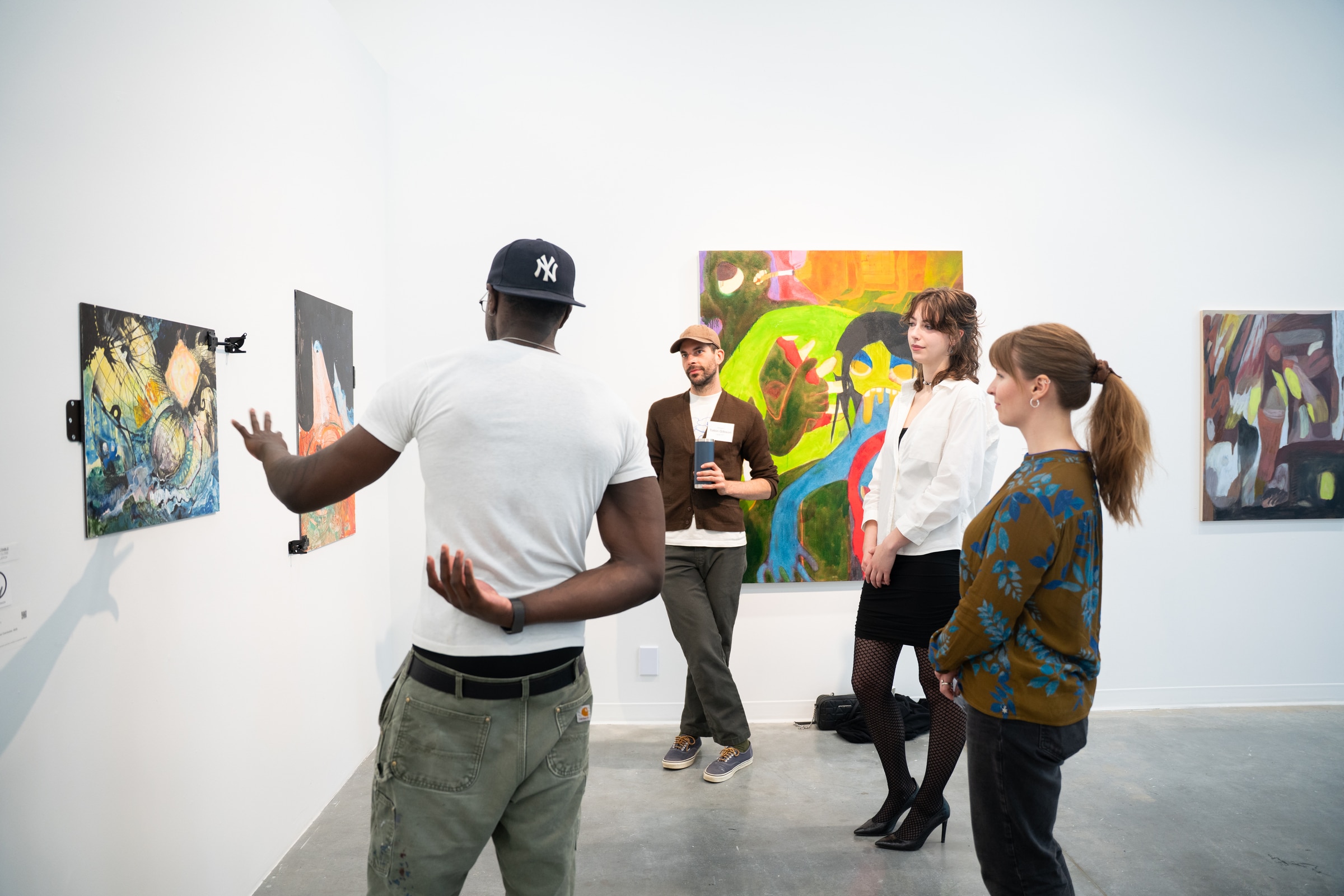 Group of students engaged in a critique session in front of colorful paintings, one student gesturing toward the work while others listen attentively.