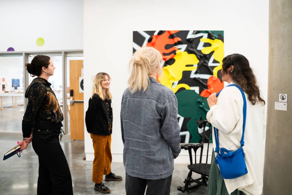 People stand with their back to the camera, discussing a large, colourful painting hanging on the exhibition space wall.