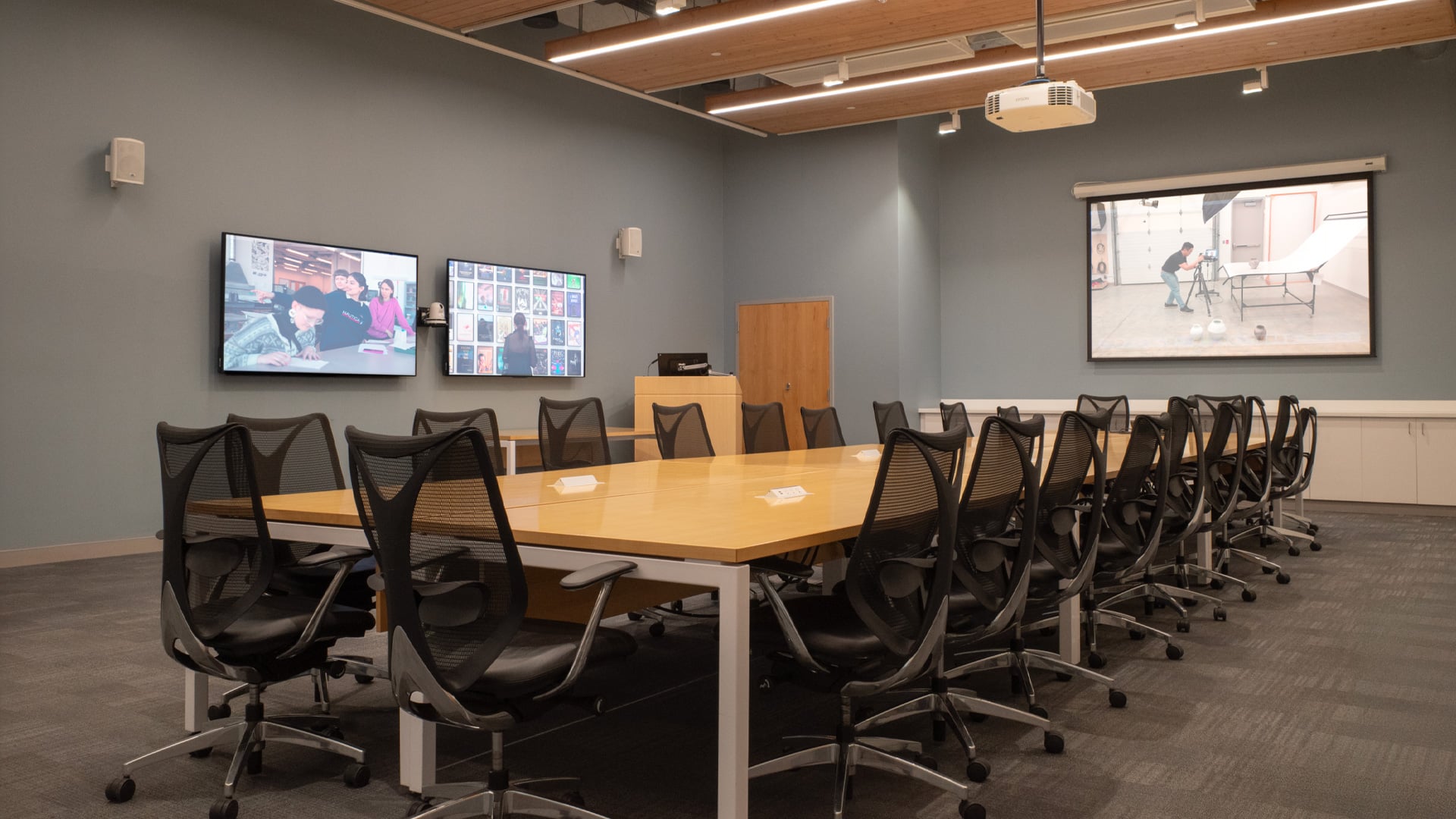 Room D2315, the Large Boardroom at Emily Carr University of Art + Design, is a modern, professionally equipped space available for rental. The room features a long wooden conference table surrounded by ergonomic black mesh chairs, ideal for meetings, workshops, or presentations. It includes advanced audiovisual technology with dual wall-mounted monitors displaying dynamic content, a large projection screen, and a ceiling-mounted projector. The neutral gray walls, warm wood accents, and ambient lighting create a versatile and inviting environment suitable for collaborative work and creative discussions.