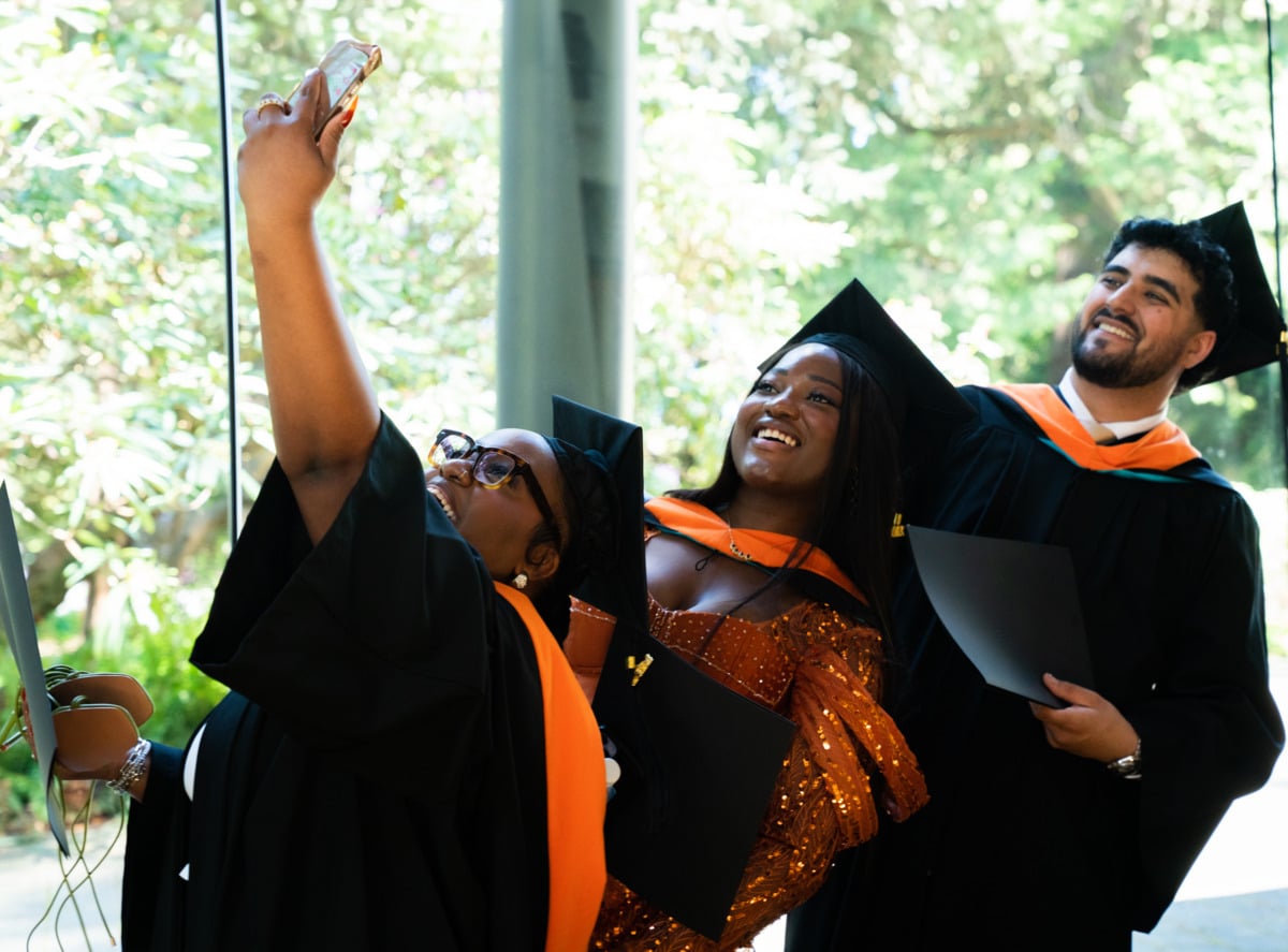 Three graduates in caps and gowns smile and pose for a selfie, celebrating together on graduation day.