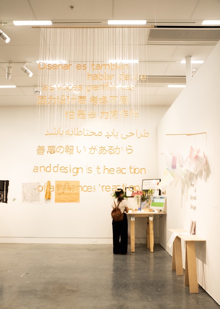 An art installation with phrases in multiple languages, including Spanish, Chinese, Arabic, Japanese, and English, made from cut-out letters suspended on strings from the ceiling. The text reads, “and design is the action of audiences’ reaction.” A visitor with a backpack stands at a wooden table beneath the text.