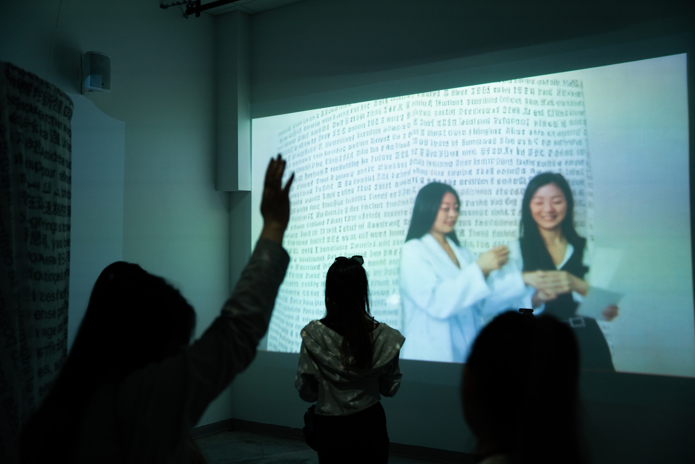 A projected video on a wall shows two women smiling and interacting in front of a backdrop filled with dense black text. In the foreground, silhouettes of viewers raise their hands toward the projection.