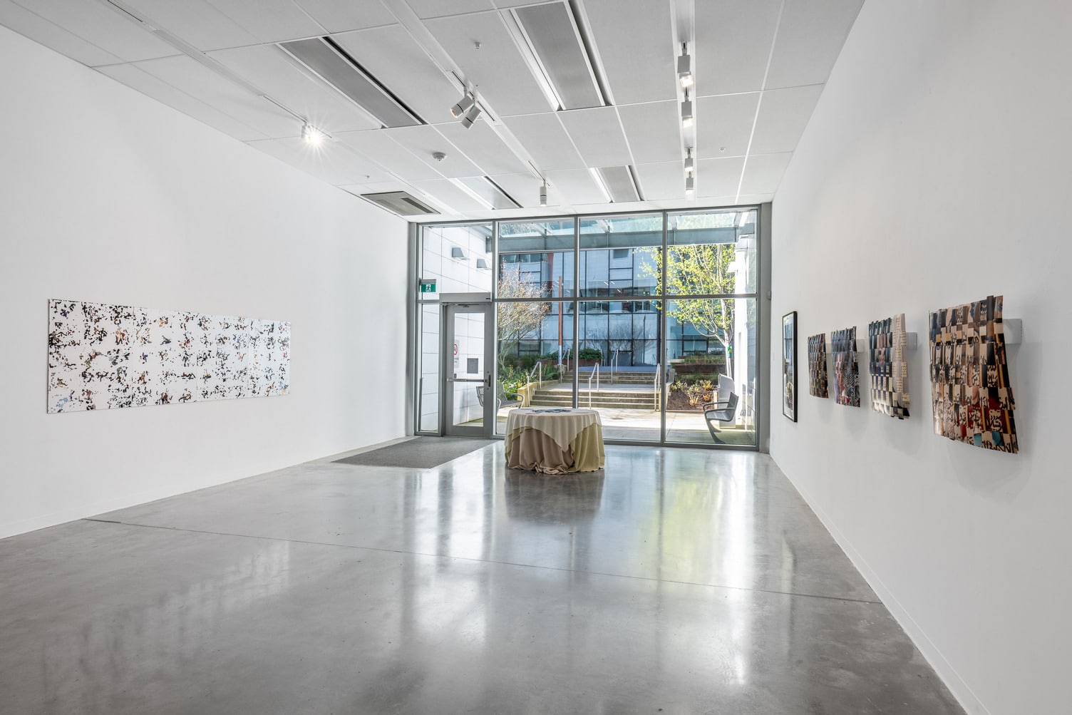 We see the Sculpture Gallery, located on Level 1 of the campus. One wall is entirely windows. There is an installation in the centre, consisting of a small round table with a tablecloth, and other work on the walls.