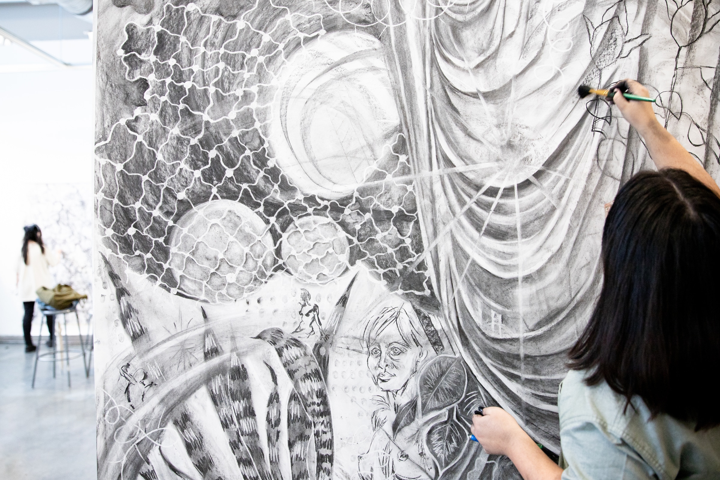 Close-up of a large charcoal drawing featuring geometric patterns, draped fabric, and a sketched human face. A person is seen in the foreground working on the artwork with a brush.