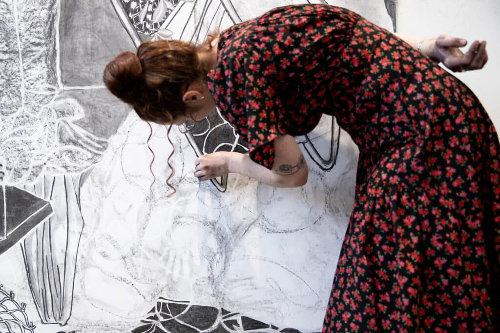 A person in a red and black floral dress bends forward, carefully working on details of a large charcoal drawing with intricate lines and textures.