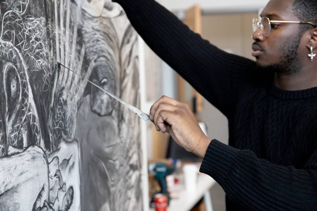 Close-up of a person in glasses and a black sweater using a drawing tool to create fine details on a large charcoal artwork featuring abstract, organic forms.