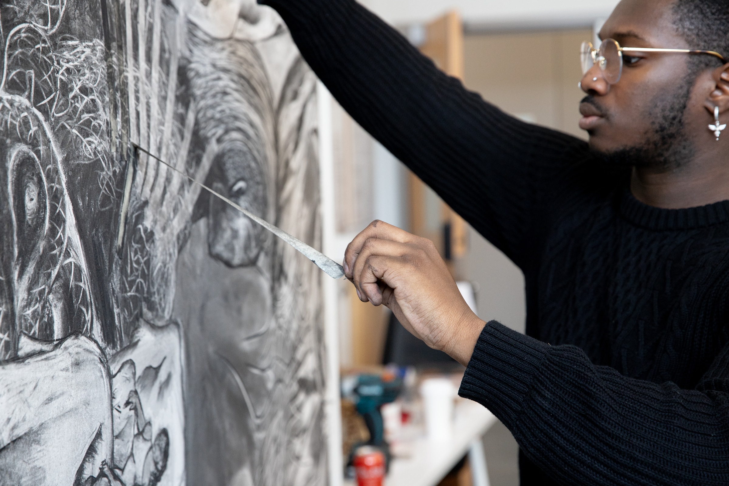 Close-up of a person in glasses and a black sweater using a drawing tool to create fine details on a large charcoal artwork featuring abstract, organic forms.