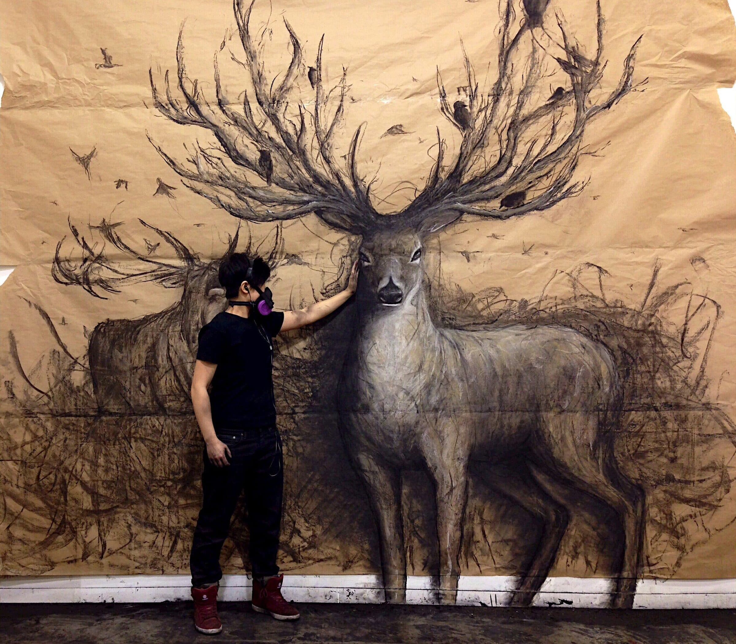 A person wearing a mask stands in front of a massive charcoal drawing on brown paper, depicting two deer with large antlers surrounded by dark, sketchy marks. The figure gently places a hand on one of the deer’s faces.