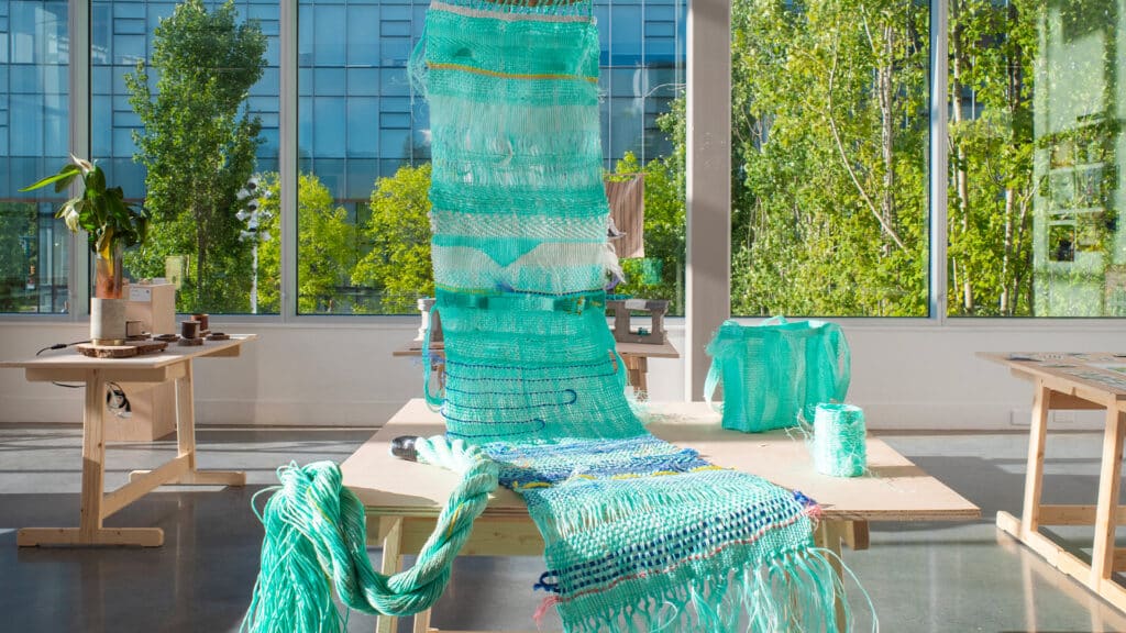 A woven installation made from bright turquoise marine plastic drapes across a wooden table in a sunlit studio, with large windows revealing green trees outside.