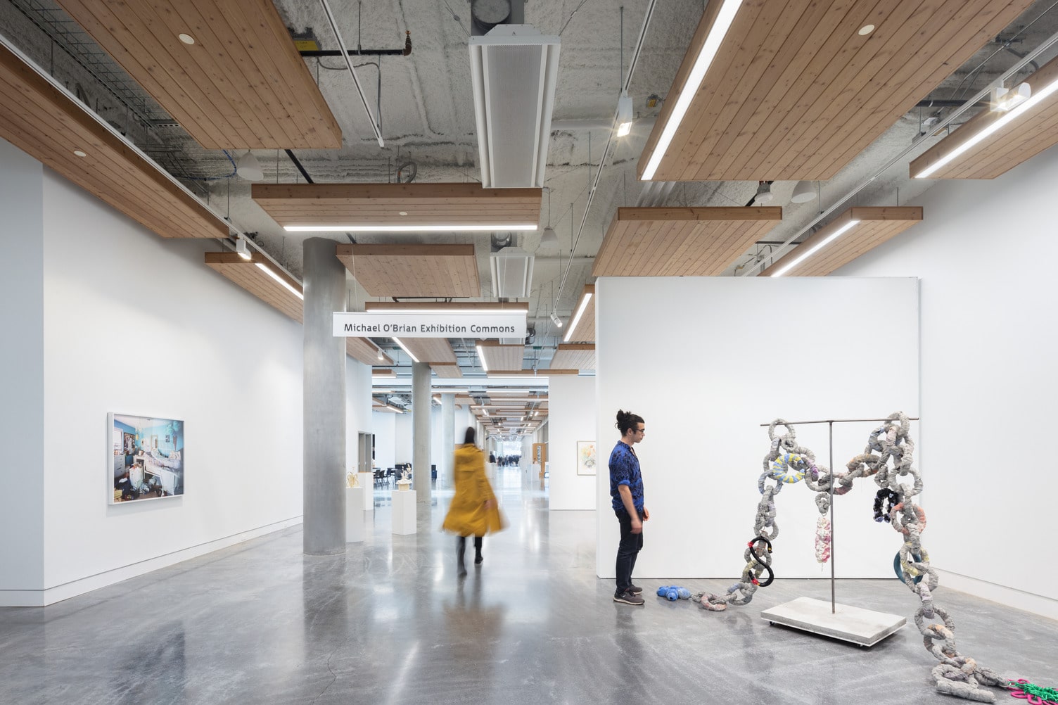 We are looking down the length of the Michael O'Brian Exhibition Commons. It is bright and airy with white walls and polished concrete floors. A large sculpture is on the right; a person looks at it while another walks down the hall.