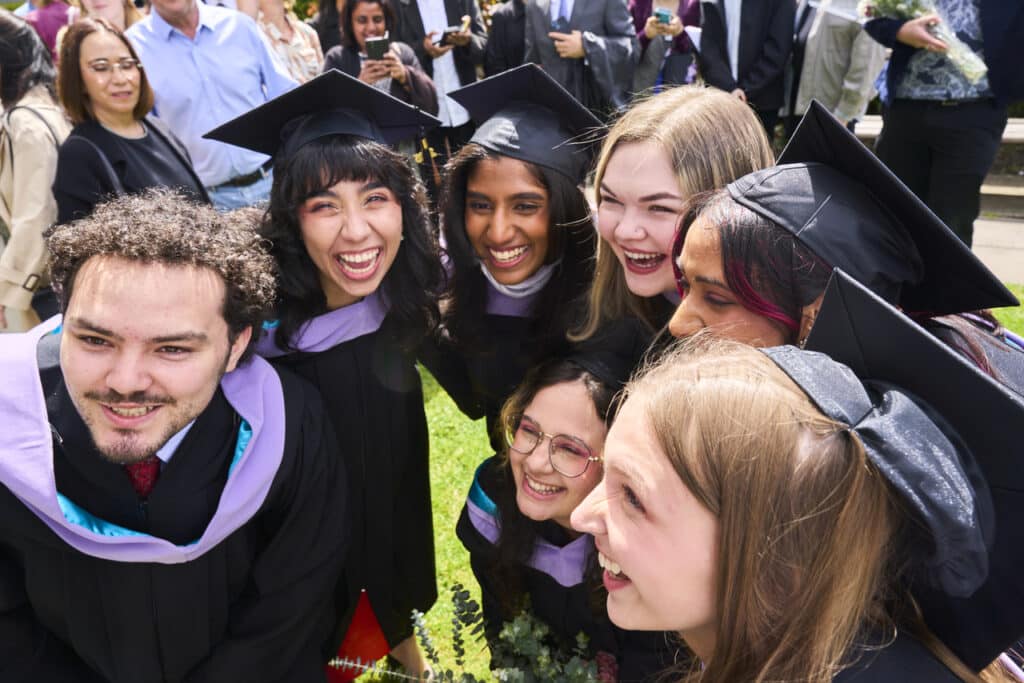 A group of seven graduates dressed in black academic gowns and caps stand closely together outdoors, smiling and laughing as they pose for a photo. They are surrounded by people in the background, some holding cameras and flowers, celebrating the occasion.