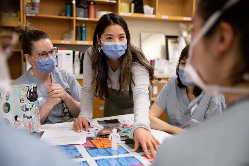 A group of healthcare workers in scrubs and face masks gather around a table with a colorful floor plan. A woman in the center, wearing a grey sweater, leans forward and points at the map while engaging others in discussion. The scene suggests teamwork and problem-solving in a hospital setting.