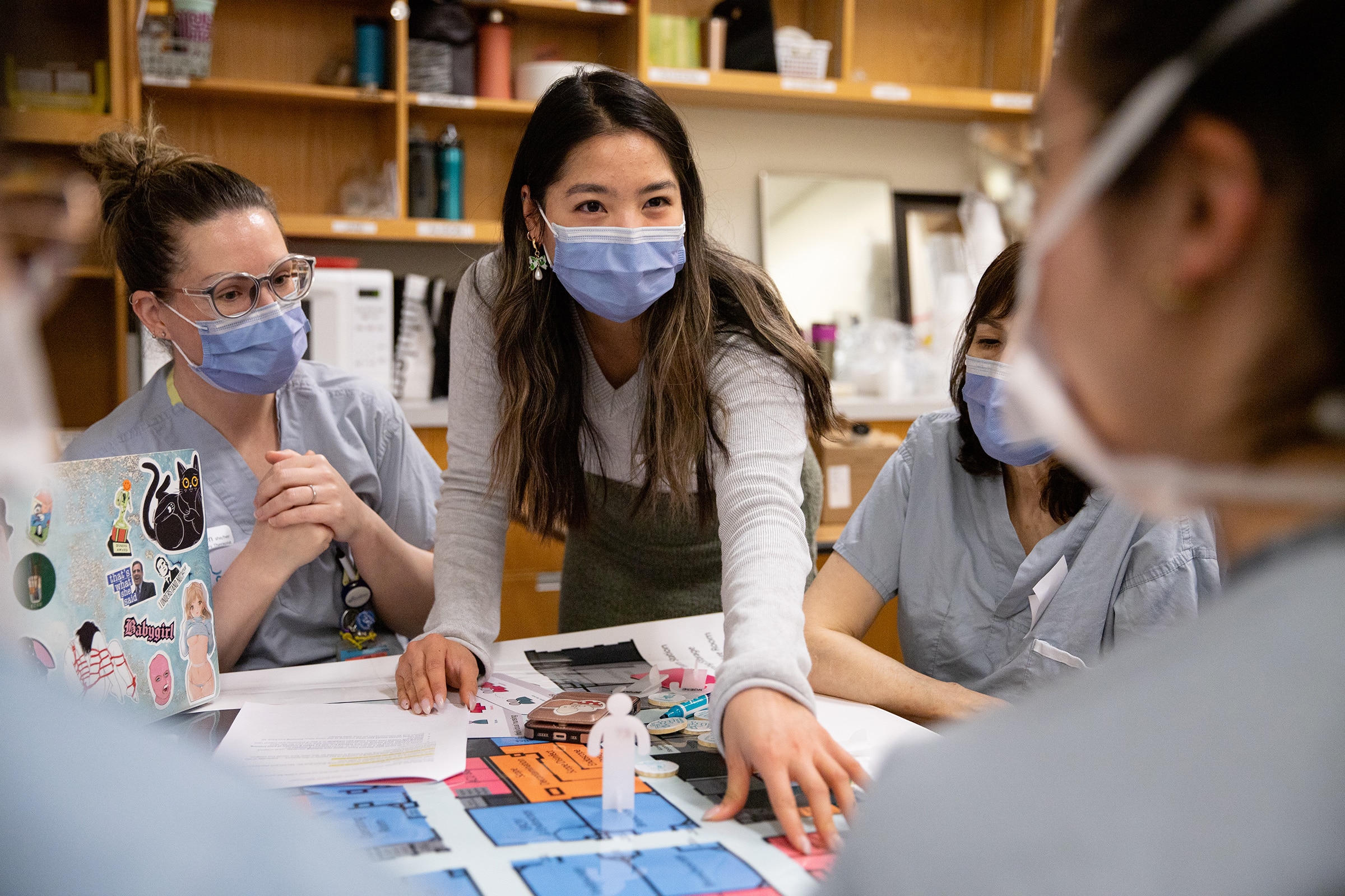 A group of healthcare workers in scrubs and face masks gather around a table with a colorful floor plan. A woman in the center, wearing a grey sweater, leans forward and points at the map while engaging others in discussion. The scene suggests teamwork and problem-solving in a hospital setting.