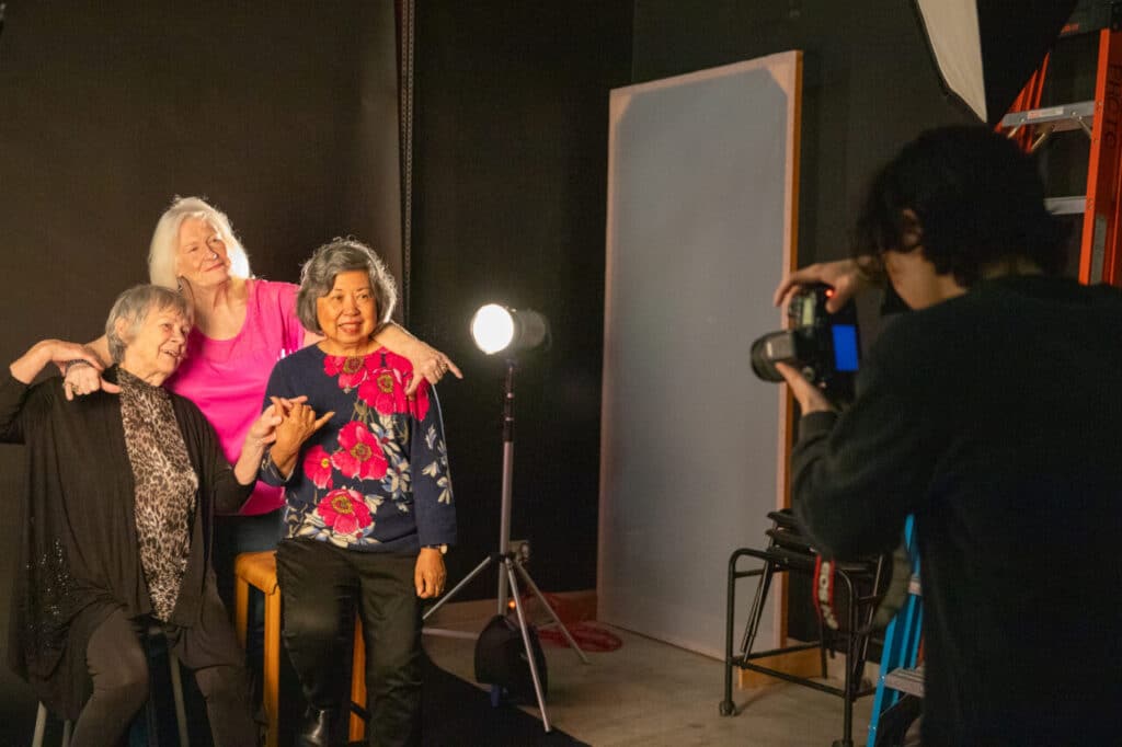 Three older people pose together in a photography studio while a photographer takes their portrait.