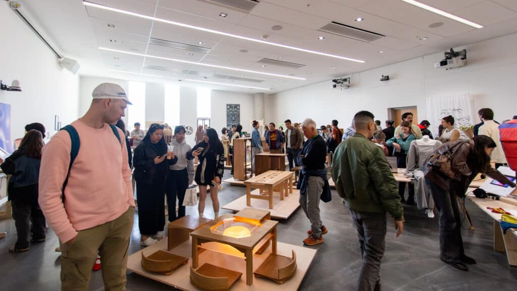 A crowded gallery space with people viewing wooden furniture pieces on display. Attendees chat, examine work closely, and take photos under bright overhead lighting.