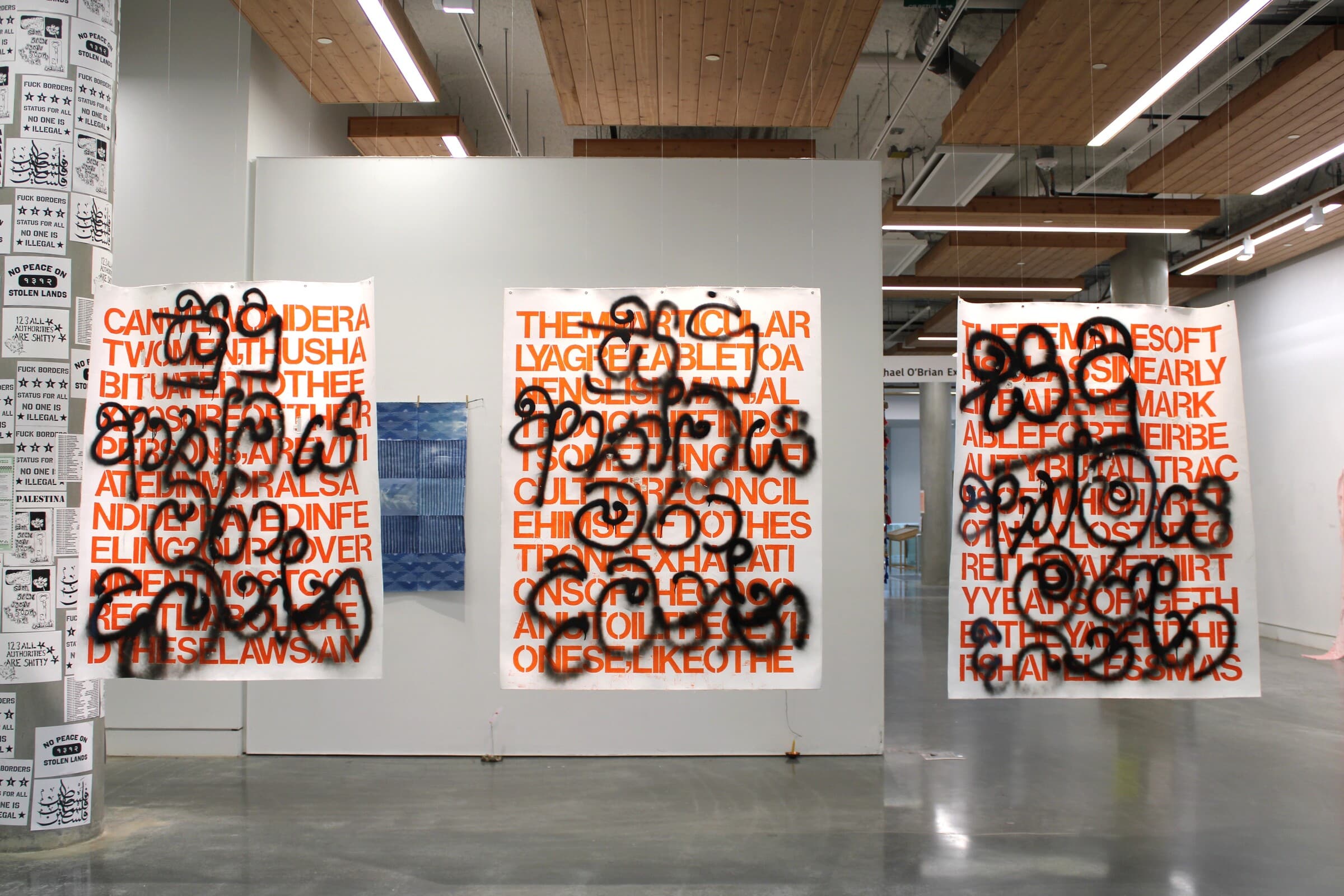 Three large posters with bold orange text hang in a row, each overlaid with heavy black spray-painted graffiti-like markings. Additional artworks, including blue fabric and posters with black text, are visible around the installation.