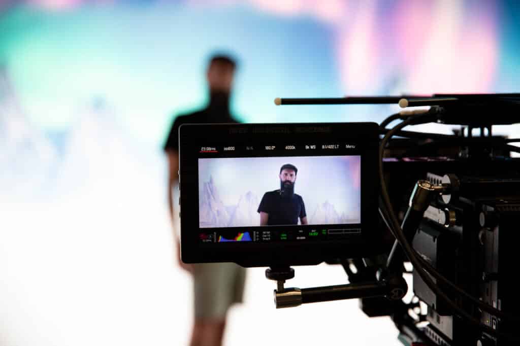 A camera monitor shows a person standing against a colorful background, captured during a video shoot in a studio.