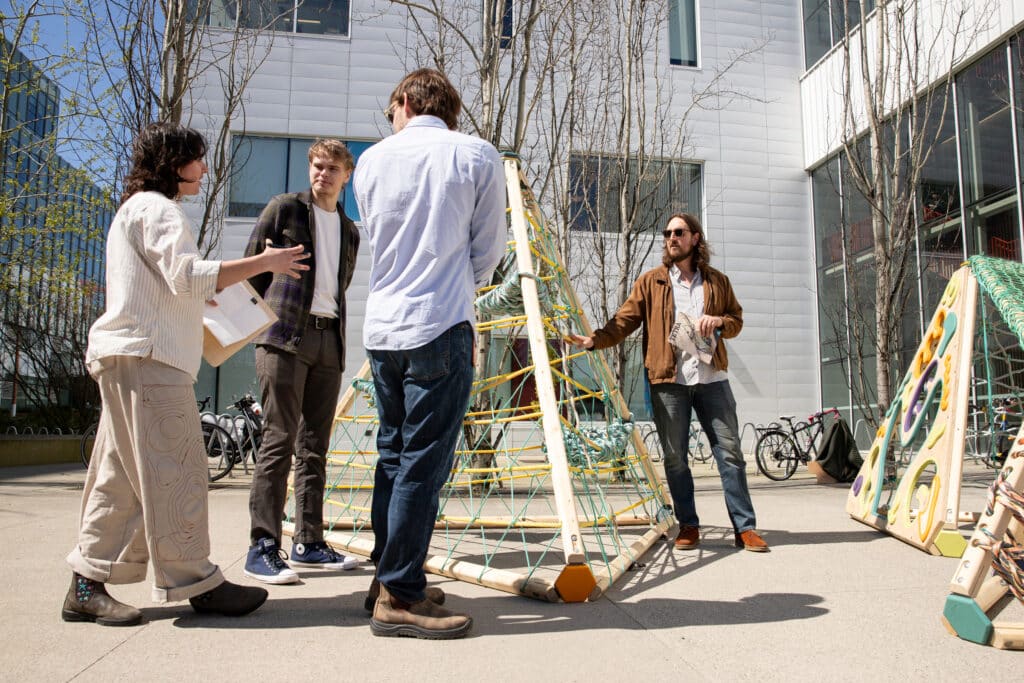 Four people gathered outdoors discussing a wooden play structure with climbing nets, set against the backdrop of a modern building.