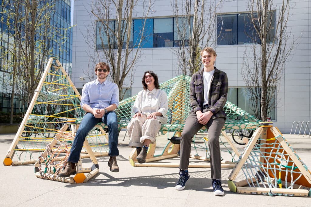 Three people sitting and smiling on a geometric wooden play structure with climbing nets, positioned outdoors in front of a modern campus building.