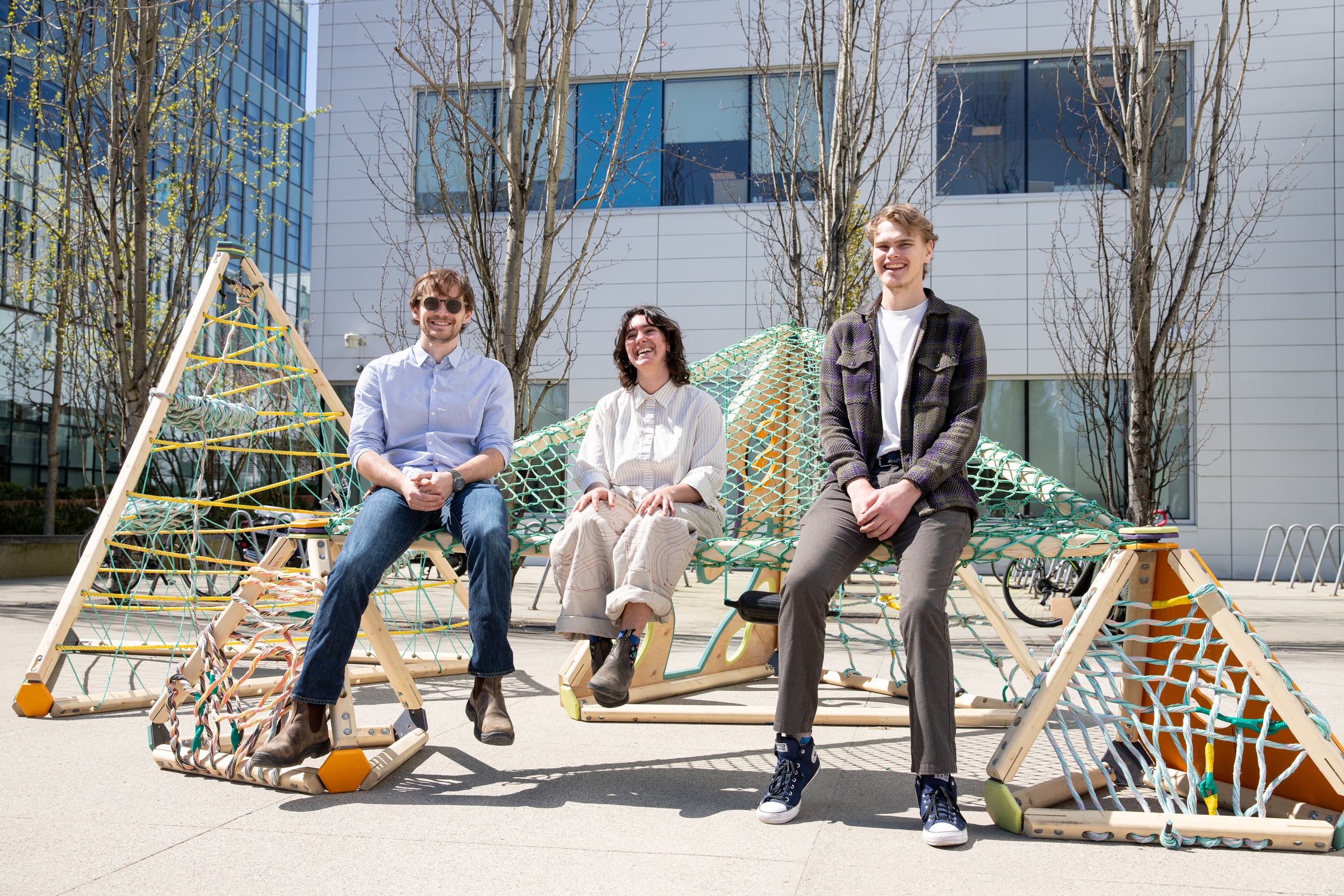 Three people sitting and smiling on a geometric wooden play structure with climbing nets, positioned outdoors in front of a modern campus building.