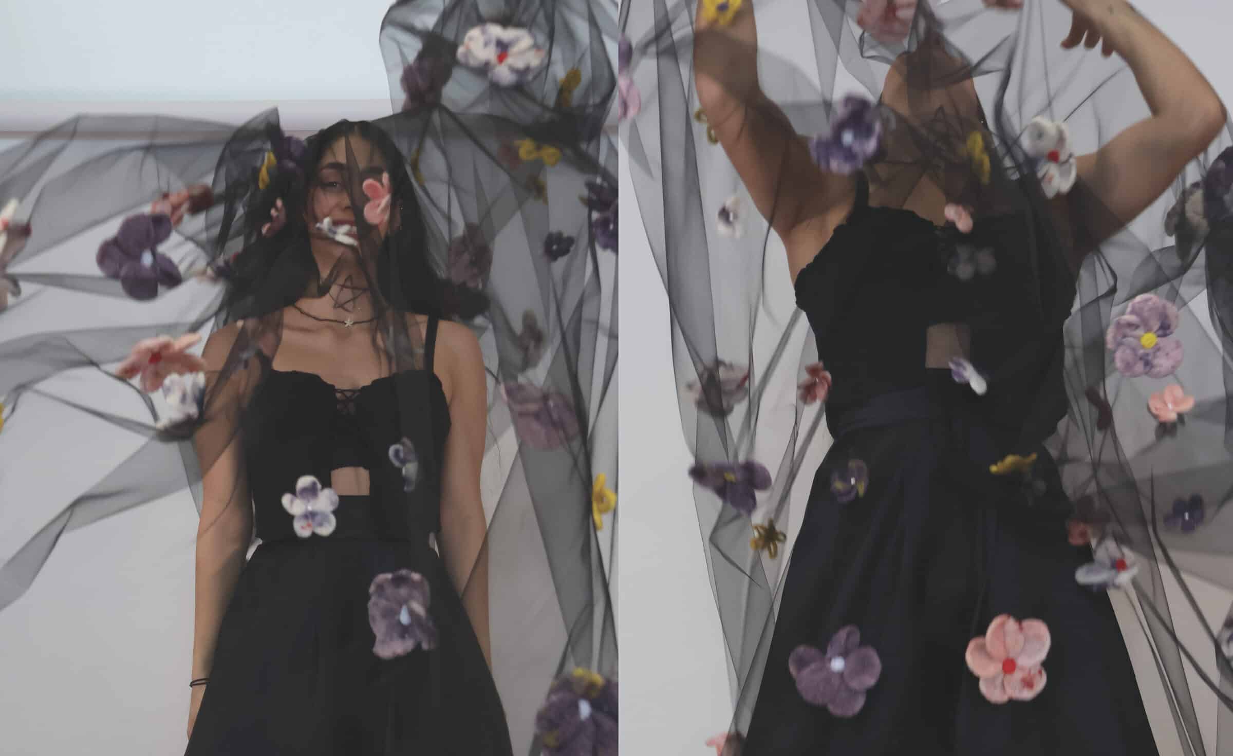 People wearing black dresses stand behind and beneath a sheer black veil decorated with colorful fabric flowers, partially obscuring their face and body.