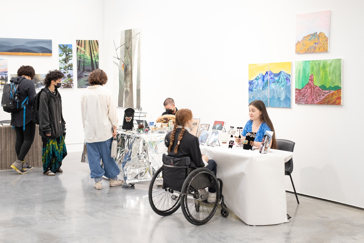 A person in a wheelchair speaks with a vendor at a table displaying jewellery and art prints during an indoor art market. Other attendees browse nearby tables, and colourful landscape paintings hang on the white gallery walls.