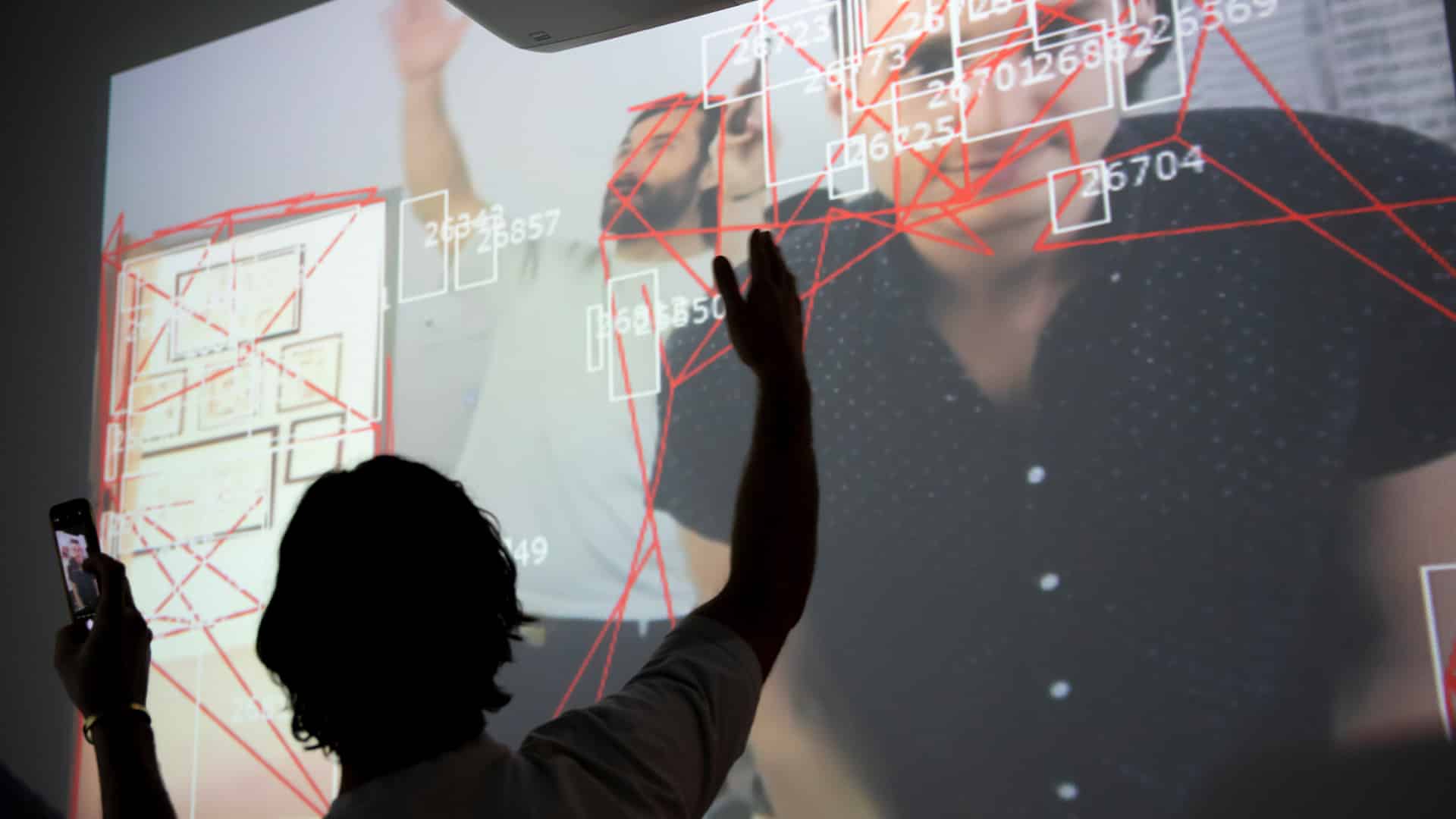 A silhouetted person interacts with a large projection showing faces, red lines and numbered boxes, while holding up a phone to capture the scene.