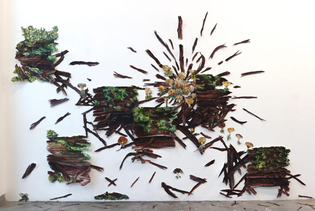 A sprawling wall installation of painted wood fragments, moss, and mushrooms arranged in bursts and clusters. The composition suggests decomposition and regrowth, with pieces radiating outward like an explosion of forest life.