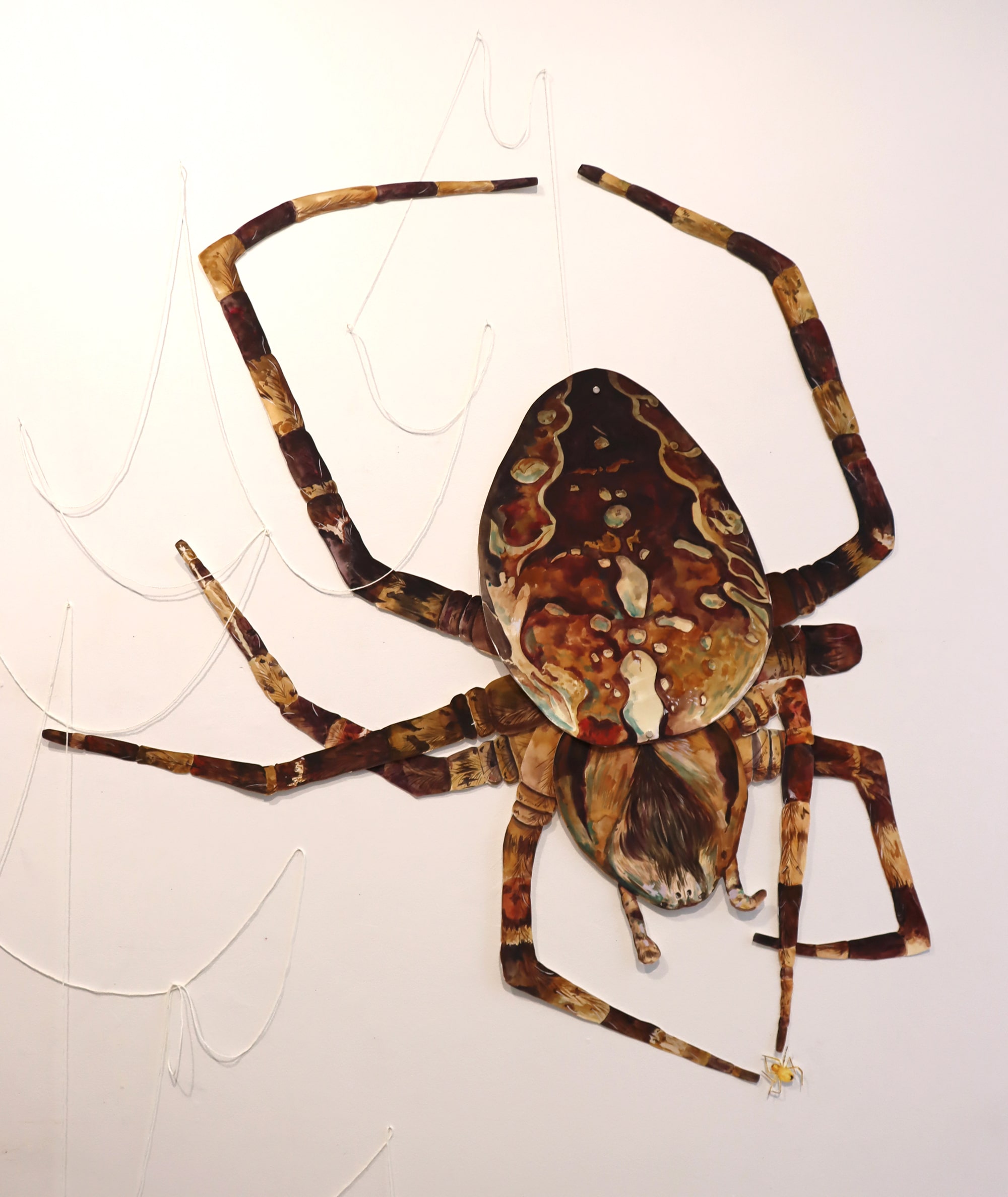 A large wall-mounted painting of a spider with mottled brown and tan legs, positioned as though spinning a delicate white web. Its body is richly textured with earthy tones, emphasizing both fragility and strength.