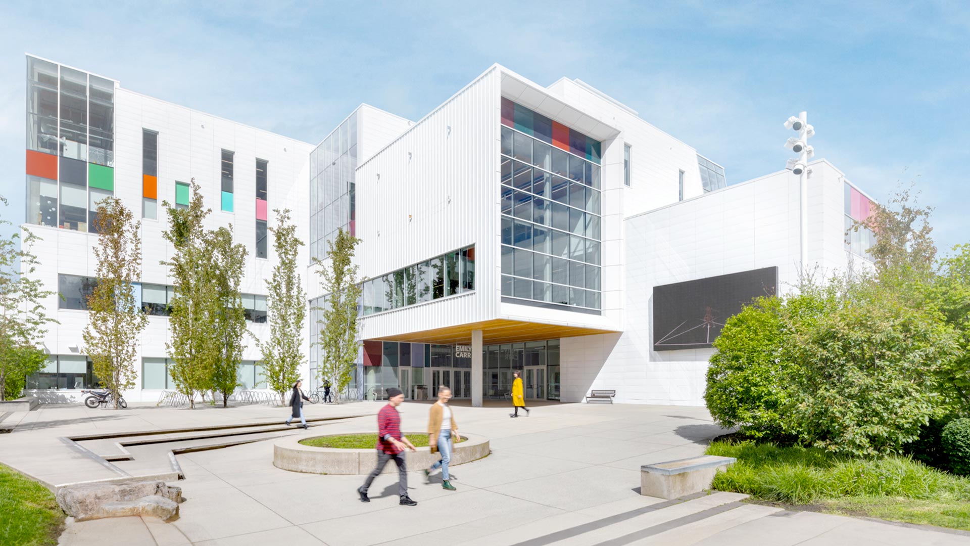 A bright, sunny day at the modern campus of Emily Carr University of Art + Design. The white, geometric building features large glass windows, colorful accent panels in red, green, and orange, and a prominent entrance with a yellow overhang. Students walk across the paved plaza, which includes landscaped planters, trees, and concrete benches. A large digital screen is mounted on the right wall, and bicycles are parked near the entrance.