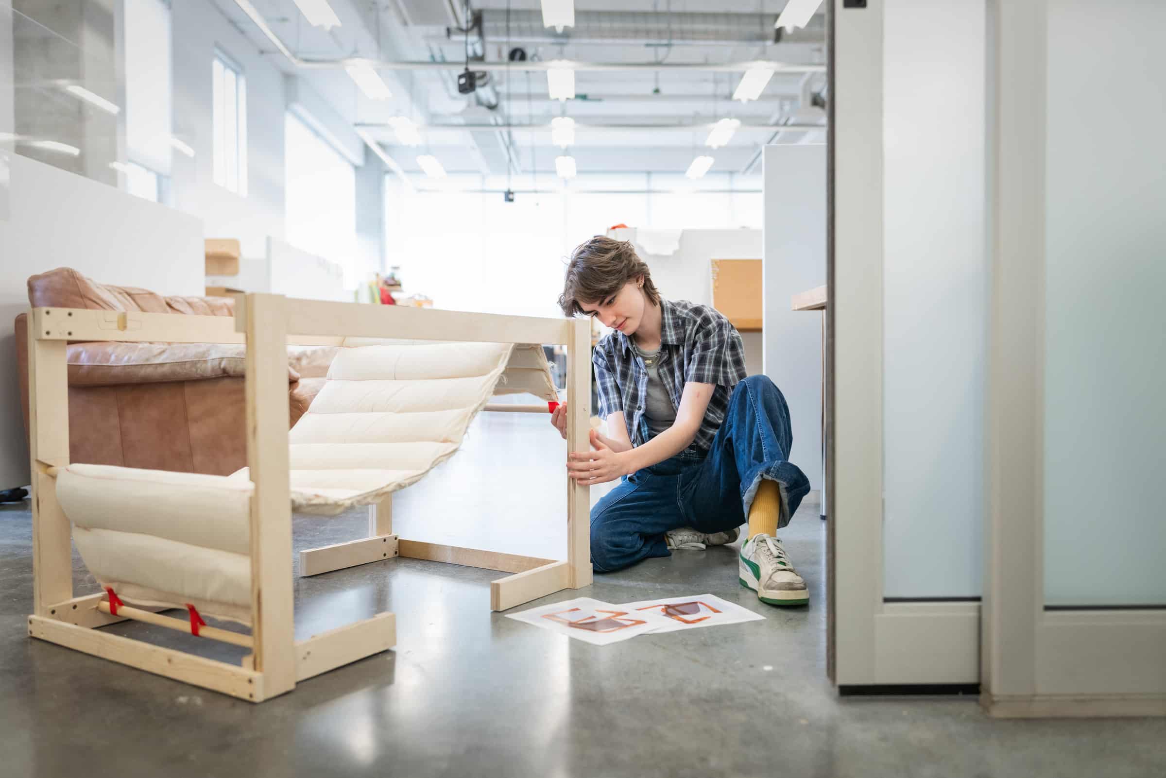A student assembling a wooden chair-like structure with a cushioned fabric seat, working on the floor of a bright studio space with design plans beside them.