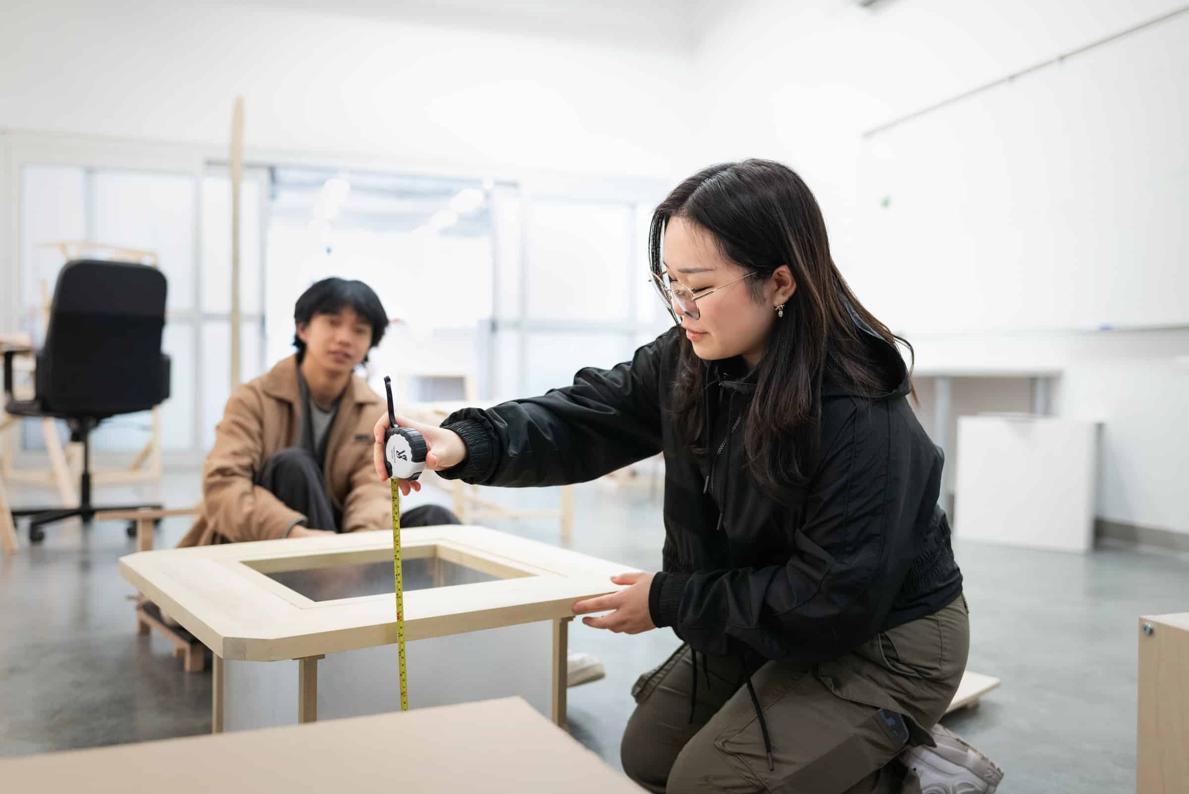 A student kneeling in a workshop and measuring a wooden furniture frame with a tape measure, while another student looks on in the background.