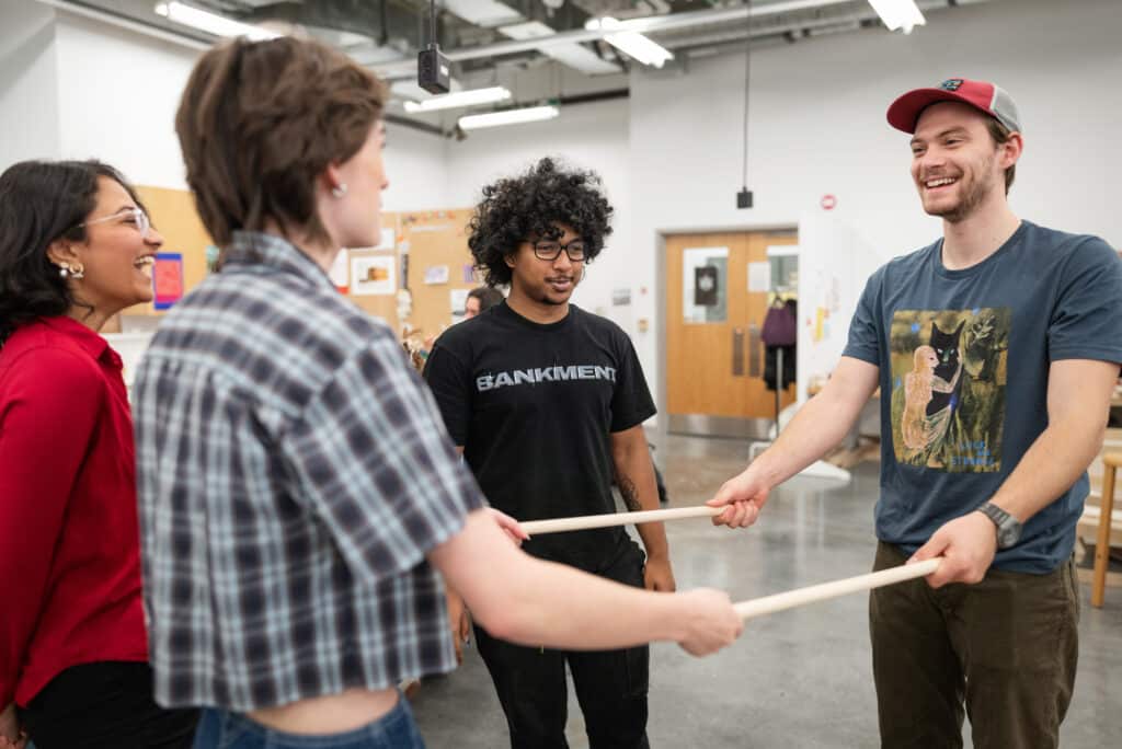 A group of four students standing in a circle in a classroom, smiling and laughing while holding two long wooden dowels between them.