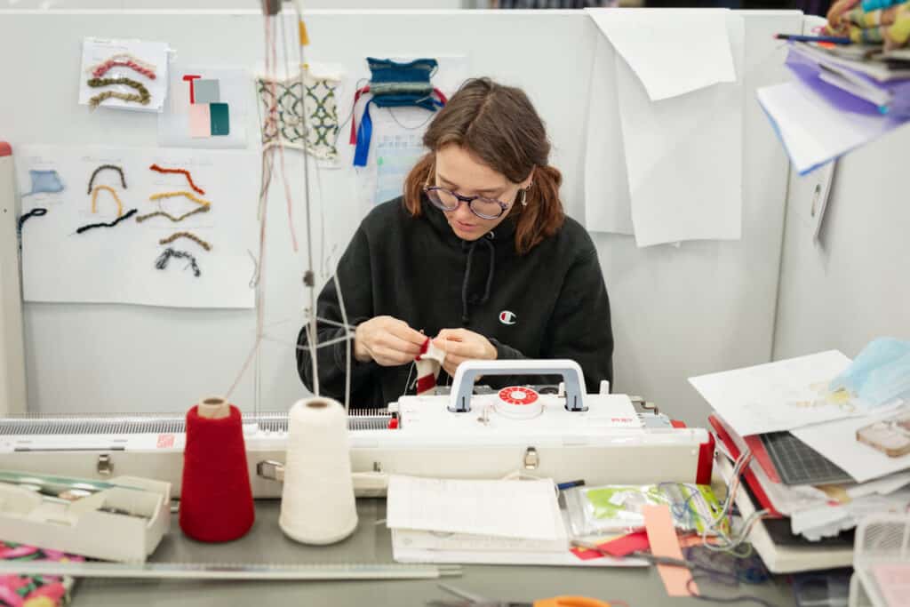 Person sits at a cluttered desk operating a knitting machine. Spools of yarn, swatches of fabric and reference notes are spread around the workspace.