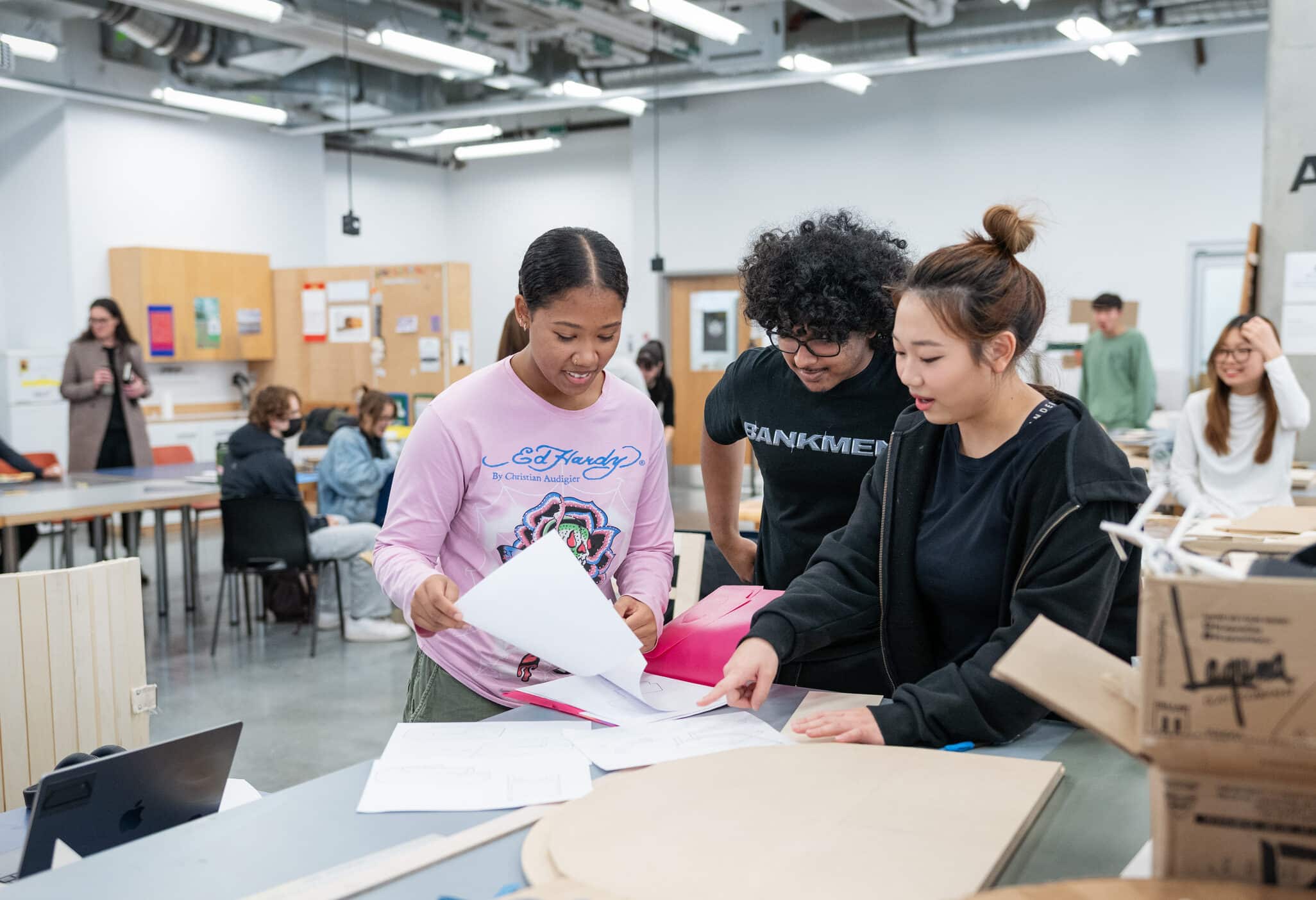 Three students standing at a work table in a classroom, reviewing papers and pointing at sketches, with other students and materials visible in the background.